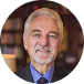 Ellipse 37 Portrait of a middle-aged man with a gray beard, wearing a blue shirt and dark blazer, in a warmly lit bookstore background.
